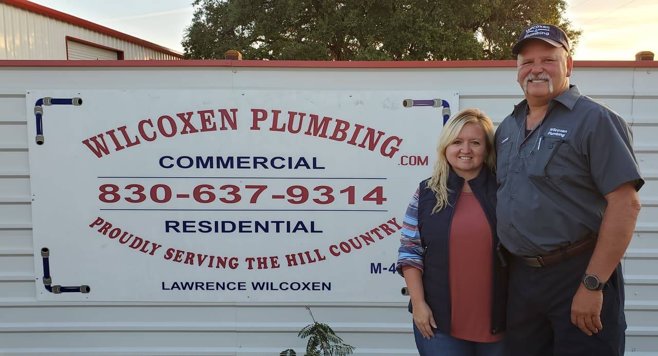 Lawrence Wilcoxen of Wilcoxen Plumbing in a work shirt and cap, standing with a smiling woman in front of the company sign
